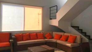 A living room with a large window, a curved sofa with orange cushions, a wicker coffee table, and a staircase with a black railing. - Tierra Linda Girardot (Girardot)