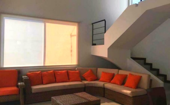 A living room with a large window, a curved sofa with orange cushions, a wicker coffee table, and a staircase with a black railing.
