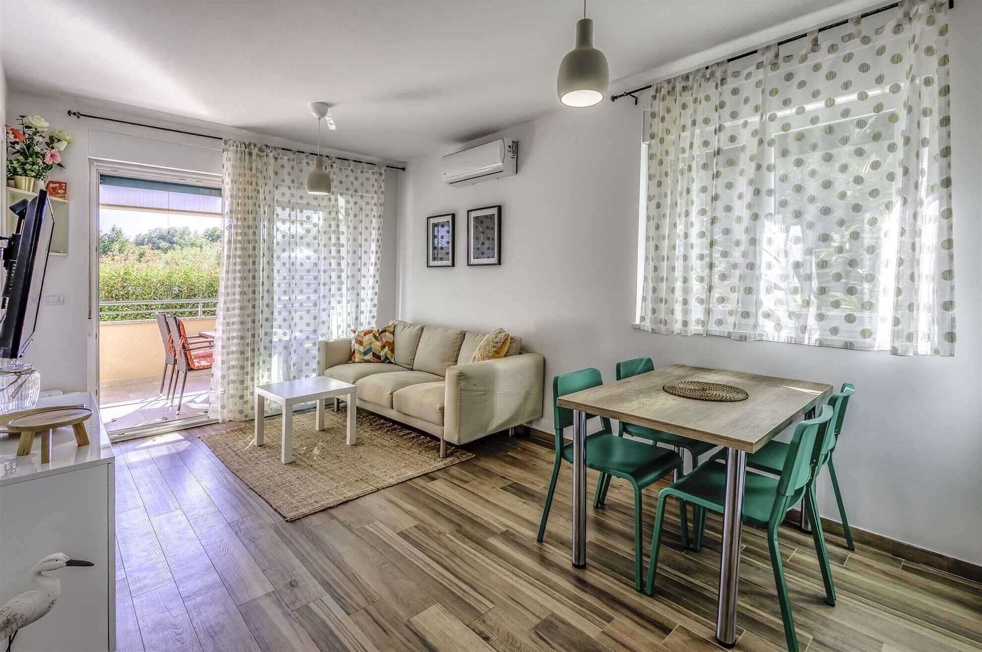 A living room with a sofa, a coffee table, a dining table with chairs, and a view of the outdoors through a door.