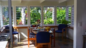 Un coin repas avec une table dressée pour quatre personnes, entourée de chaises, de grandes fenêtres donnant sur des plantes tropicales, et un espace cuisine situé à gauche.