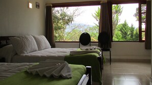 A hotel room with two beds, a chair, and large windows offering a view of greenery and mountains.