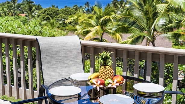 Balcón con mesa dispuesta para comer, con una piña y otras frutas, con vista a un paisaje tropical de palmeras y el océano.