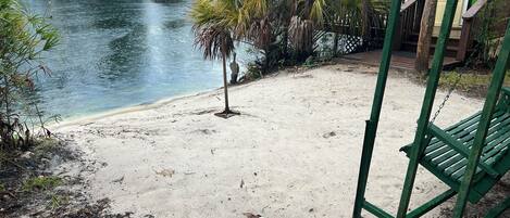 A sandy beach area with a small palm tree, a swing set, and a boat in the background.