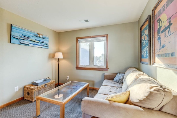 Un salon avec un canapé beige, une table basse en verre, un panier en osier, une lampe et un tableau représentant des bateaux accroché au mur.