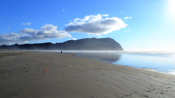Una extensa playa de arena con un mar tranquilo, una montaña distante y algunas personas caminando.