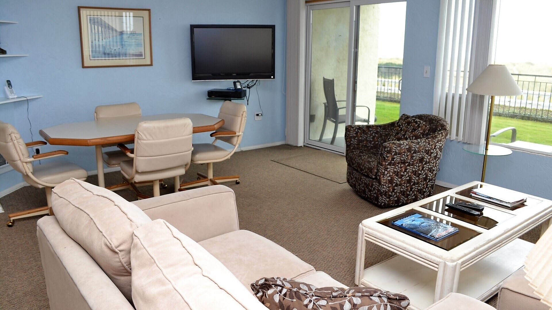 A living room with a sofa, a patterned armchair, a television, a table with books and remotes, and a view of the outdoors.