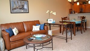 A living room with a brown leather sofa, a glass coffee table, and a dining area with a wooden table and chairs. - 2Bed/2Bath Upscale, River Front Condos. Close to Beach & Convention Ctr (Seaside)