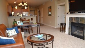 A living room with a brown leather sofa, a glass coffee table, a fireplace, and a kitchen area in the background. - 2Bed/2Bath Upscale, River Front Condos. Close to Beach & Convention Ctr (Seaside)