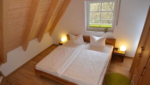 A bedroom with a wooden bed, two bedside tables with lamps, a window with a view of trees, and a green rug on the floor. - Vacation apartment North Sea (OG) (Sellin)