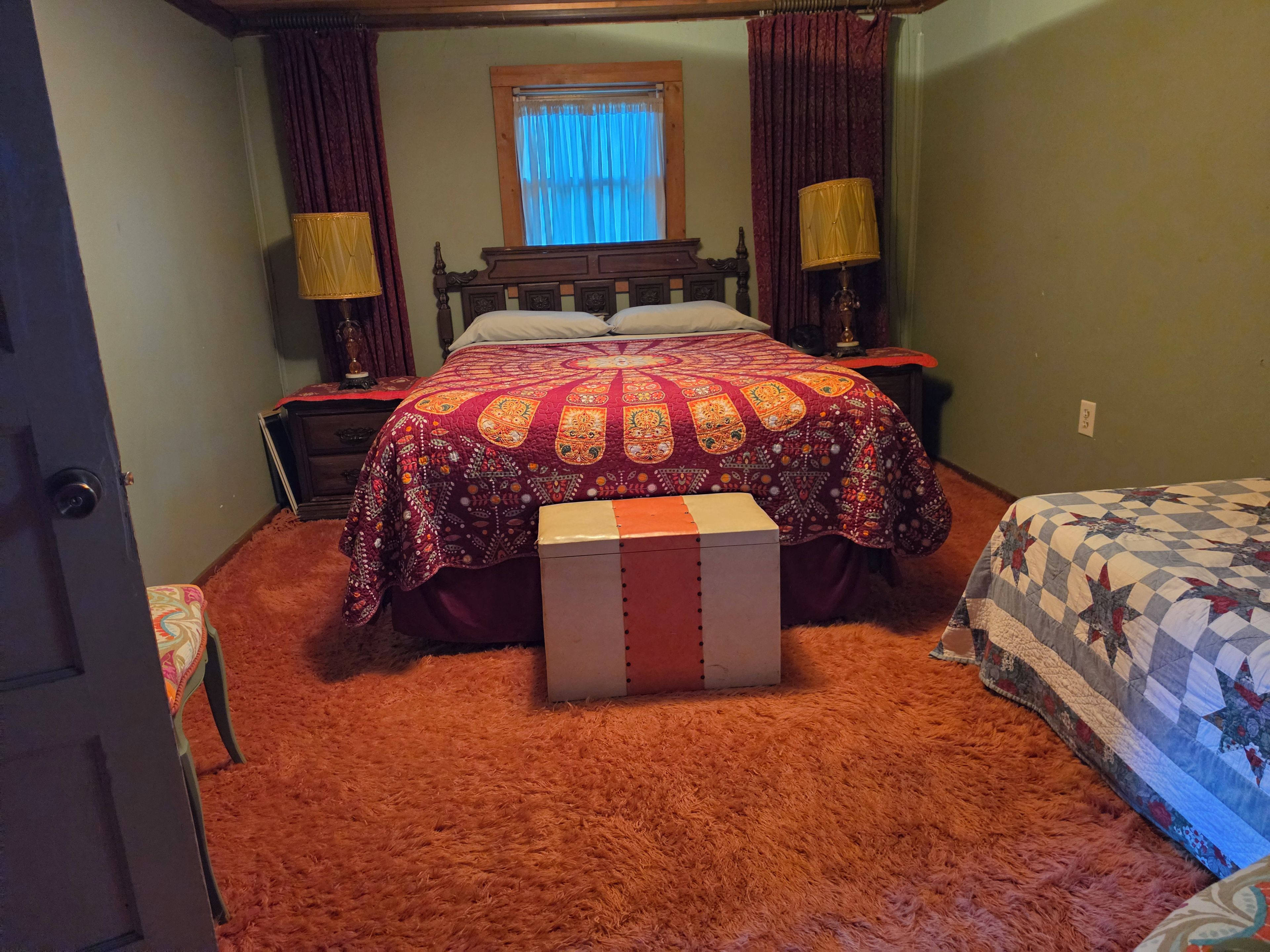 Un dormitorio con una cama grande, dos mesitas de noche, una ventana con cortinas y piso alfombrado.