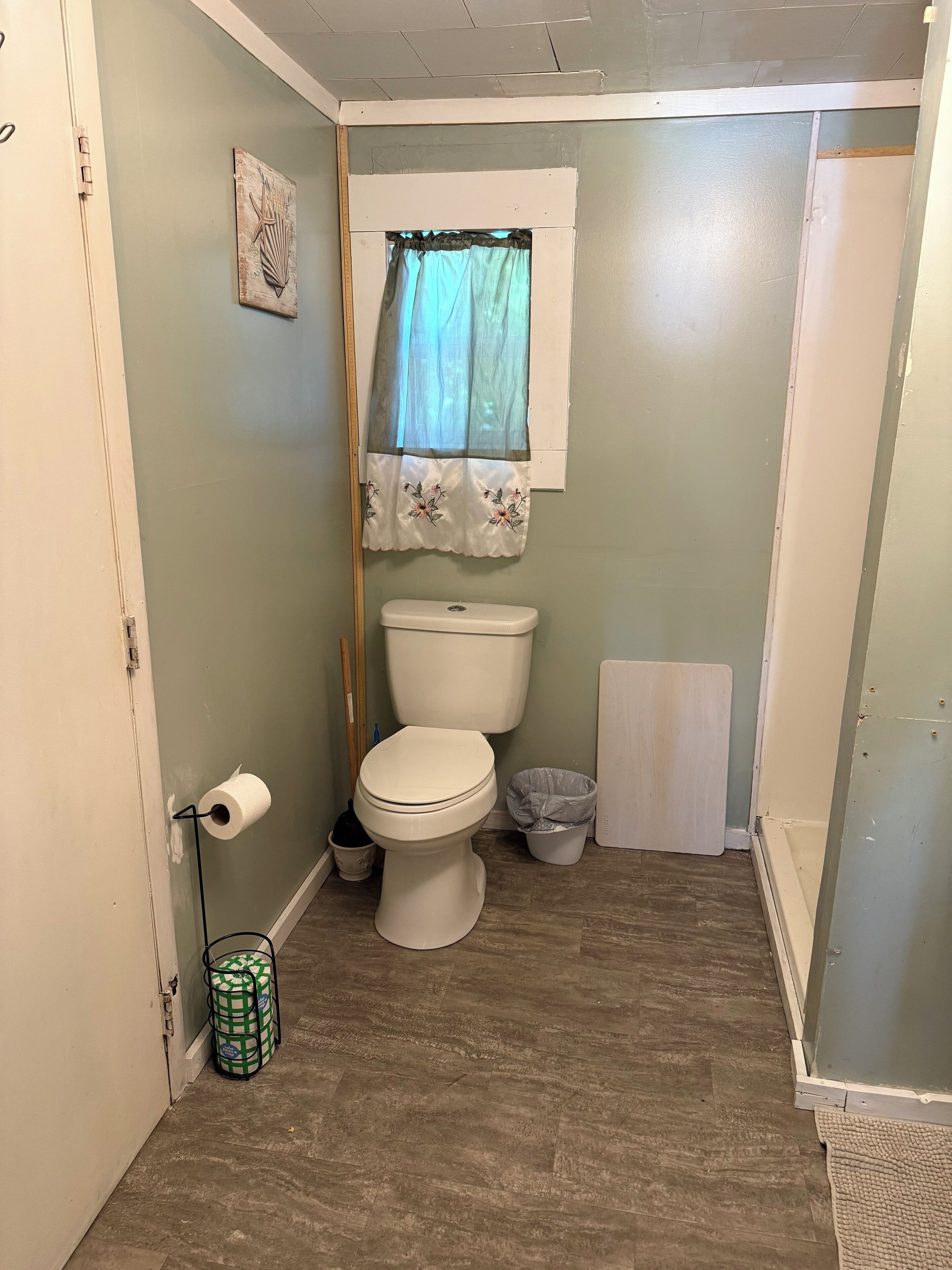 Een kleine badkamer met een toilet, een prullenbak en een raam met een dunne gordijn.