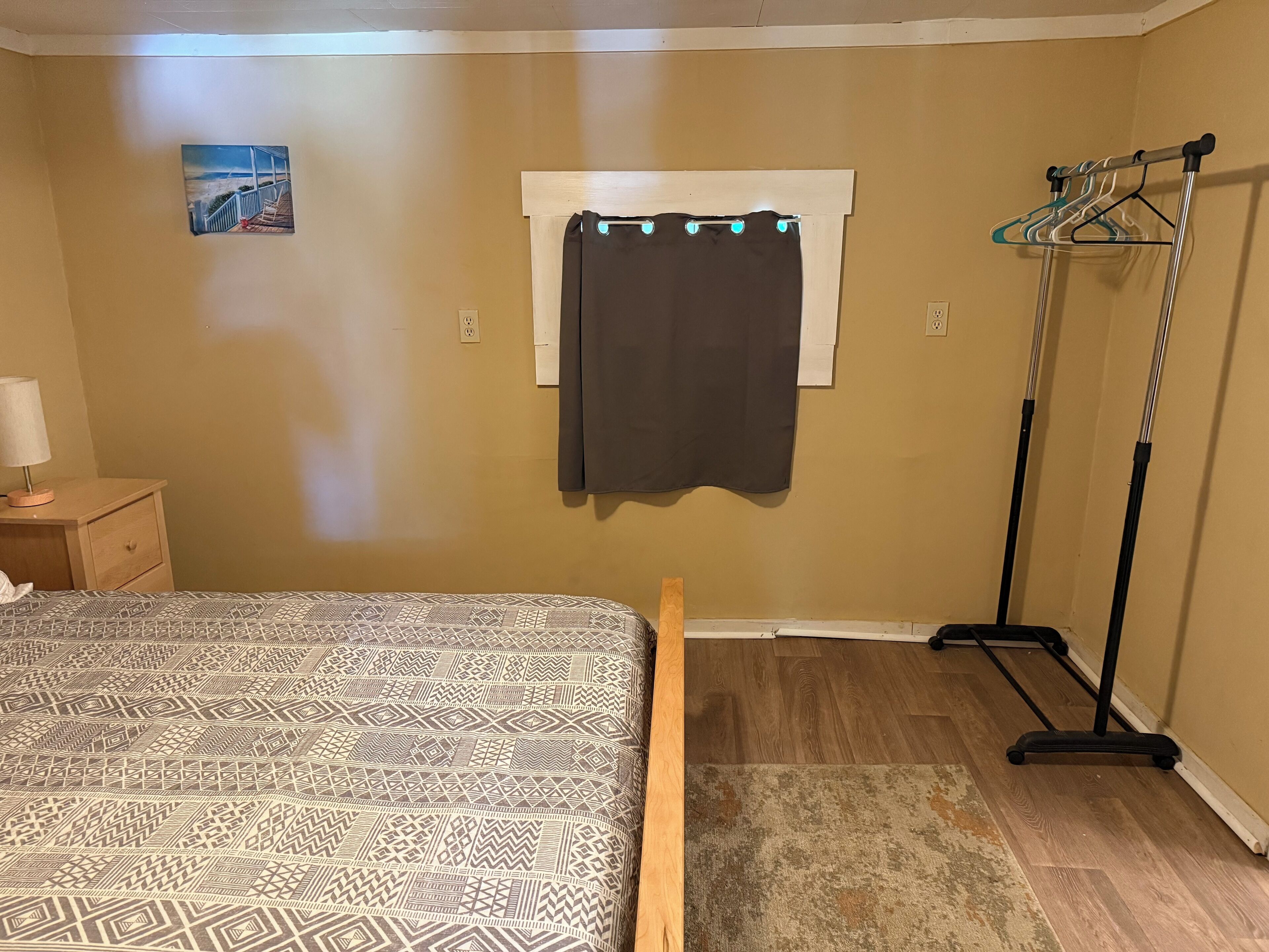A bedroom with a bed, a nightstand, a window with a curtain, and a clothing rack.
