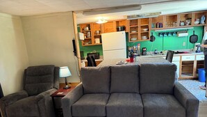 A living room with a grey sofa, a grey armchair, and a small table with a lamp. A kitchen is visible in the background with wooden cabinets and various kitchen items. - Rustic intown country apartment on the Moosehead trail (Sangerville)