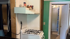 A compact kitchen with a gas stove, a small shelf with spices, and a dining area with a checkered tablecloth. - Rustic intown country apartment on the Moosehead trail (Sangerville)