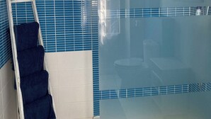 Kylpyhuone, jossa on lasiseinäinen suihkukaappi, valkoisilla ja sinisillä laatoilla päällystetty seinä sekä valkoinen pyyhekoukku, jossa on pyyhkeitä.