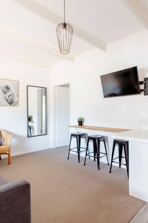 A modern living room with a flat-screen TV, a dining area with stools, and a large mirror.
