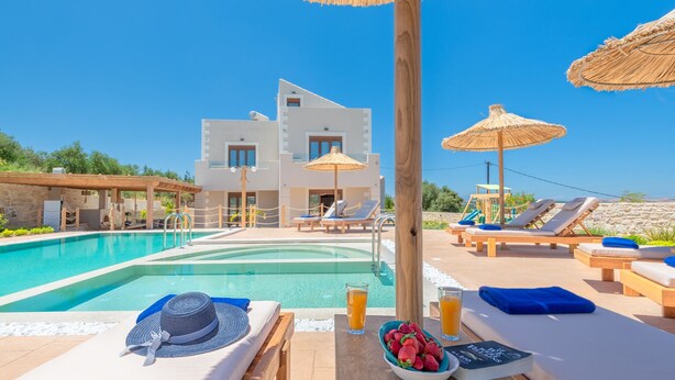 A poolside area with lounge chairs, umbrellas, and a clear view of a modern building in the background. - Ivoni Villa, Spacious Family Retreat • Pool & Play (Perama)