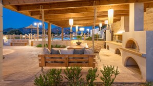An outdoor seating area with wooden benches and tables, a pool, and a stone wall with an arched entrance. - Ivoni Villa, Spacious Family Retreat • Pool & Play (Perama)