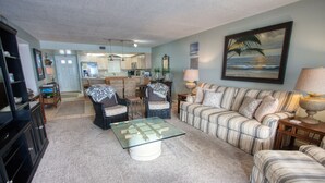 A living room with a sofa, armchairs, a glass coffee table, and a dining area with a kitchen in the background. - Location, location, location.  Keep a close eye on the kids pool side, from this 2nd floor 2/2 condo (New Smyrna Beach)