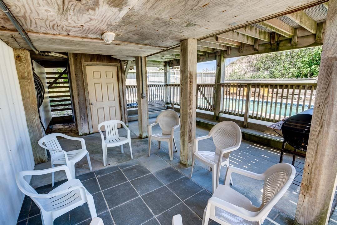 A covered patio area with white plastic chairs, a wooden door, and a view of a swimming pool.