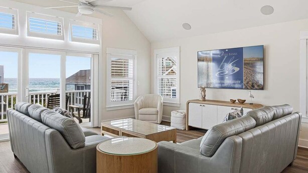 A spacious living room with a view of the ocean, a ceiling fan, and a mounted television. - Sandpiper Shore | 75 ft to the Beach | Brand New Construction (Nags Head)