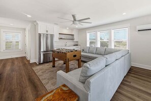 A modern living room with a grey sofa, a foosball table, a ceiling fan, and a kitchen area with white cabinets and a stainless steel refrigerator.