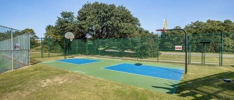 Een basketbalveld buiten, met twee blauwe velden, omgeven door een groene hekken en bomen op de achtergrond.