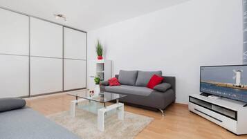 Ett modernt vardagsrum med en grå soffa, ett soffbord i glas och en platt-TV placerad på ett vitt TV-bord.