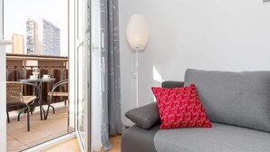 En moderne stue med en grå sofa, en rød pude med mønster og en hvid gulvlampe. En glasdør fører til et udendørs opholdsområde med fletstole og et bord.