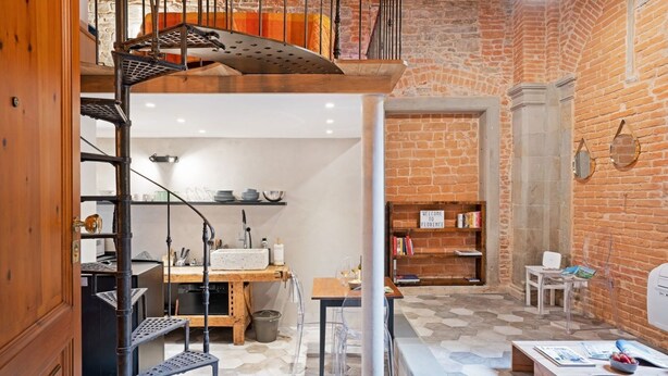Un espace de vie de style « loft », avec un mur en briques, un escalier en bois, un coin cuisine et un espace détente comprenant une table et des chaises.