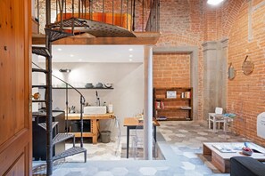 A loft-style room with a brick wall, a wooden staircase, a kitchen area, and a seating area with a table and chairs.