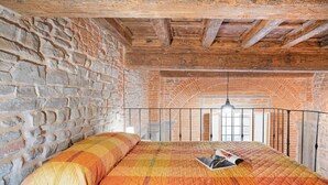 Une chambre avec un mur en briques, des poutres en bois au plafond, un lit recouvert d’une courtepointe et un luminaire suspendu.