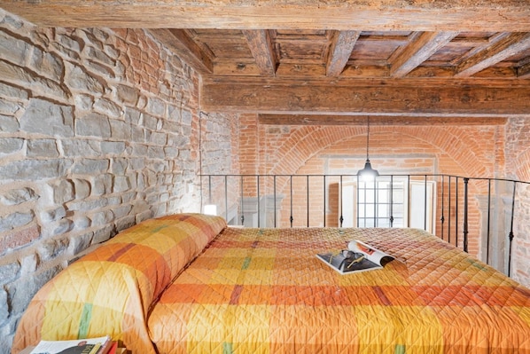 Une chambre avec un mur en briques, des poutres en bois au plafond, un lit recouvert d’une courtepointe et un luminaire suspendu.