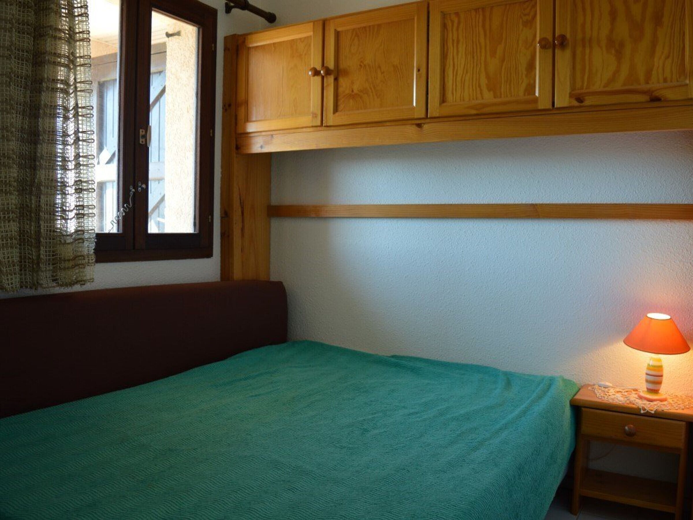 A bedroom with a bed, a nightstand, a lamp, wooden cabinets, and a window with curtains.
