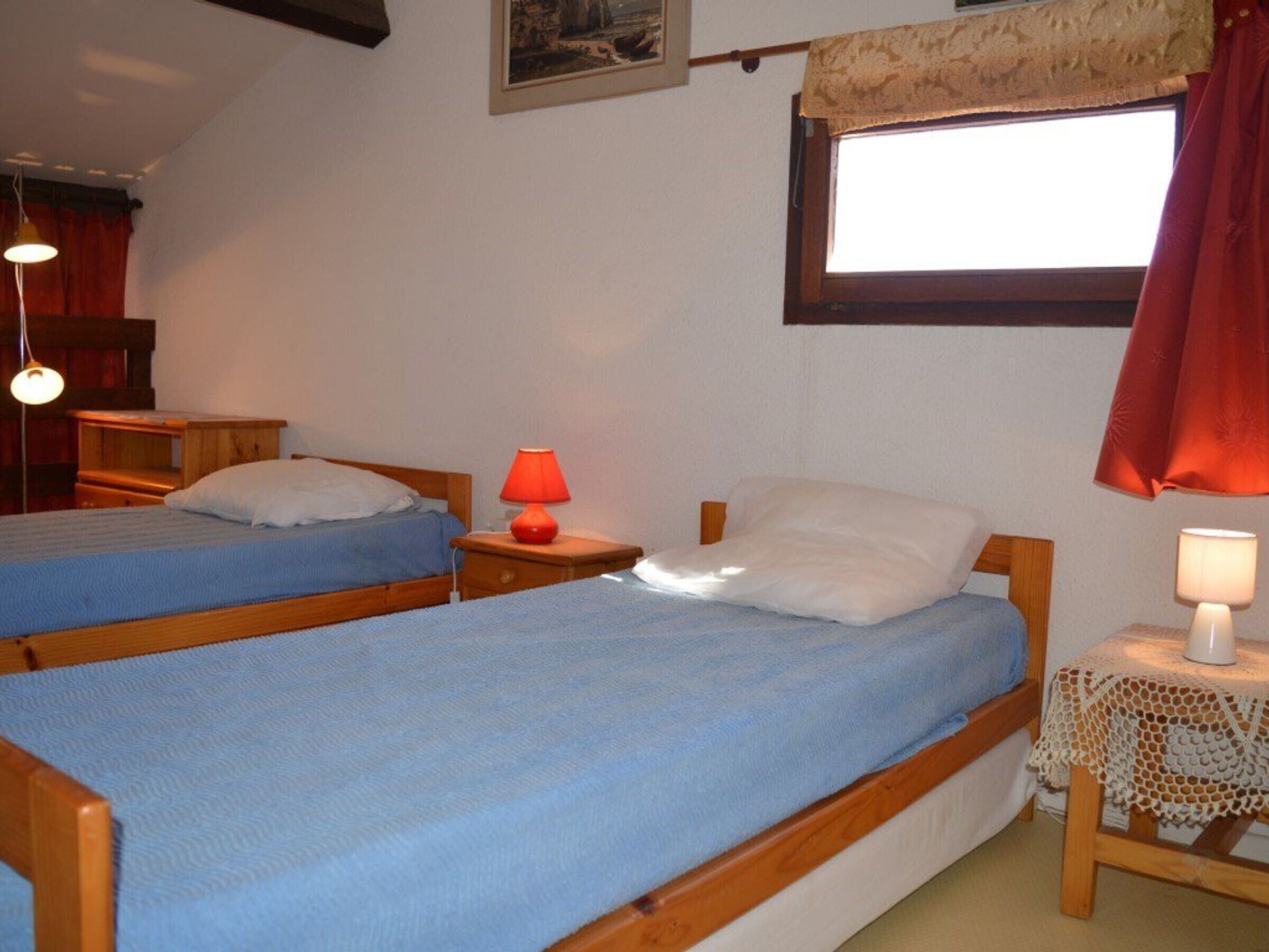Un dormitorio con dos camas individuales, una ventana con cortina, una mesita con lámpara y un cuadro colgado en la pared.