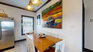 A dining area with a wooden table, white chairs, a stainless steel refrigerator, and a colorful painting on the wall.