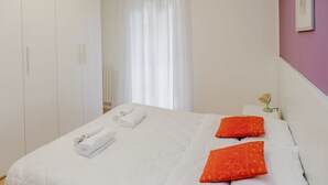 A hotel room with a bed, white curtains, a white wardrobe, and a bedside table with a lamp.