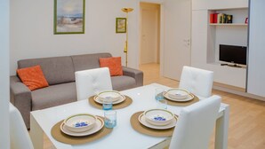A dining area with a white table set for four, a grey sofa with orange cushions, a bookshelf with books and a TV, and a framed picture on the wall.