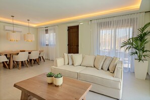 A modern living room with a beige sofa, a wooden coffee table with plants, a dining area with a wooden table and chairs, and a large window with sheer curtains.