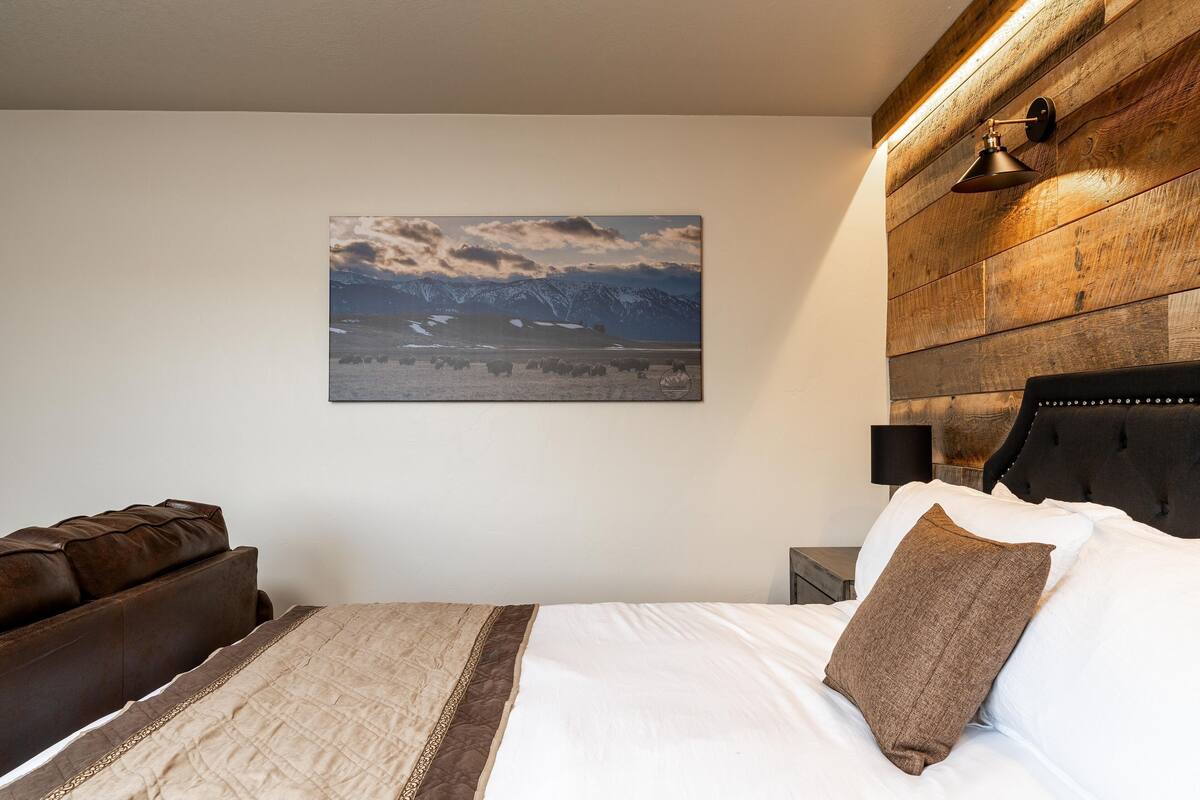 A bedroom with a bed, a brown leather sofa, a wooden headboard, a mounted picture of a landscape, and a wall-mounted lamp.