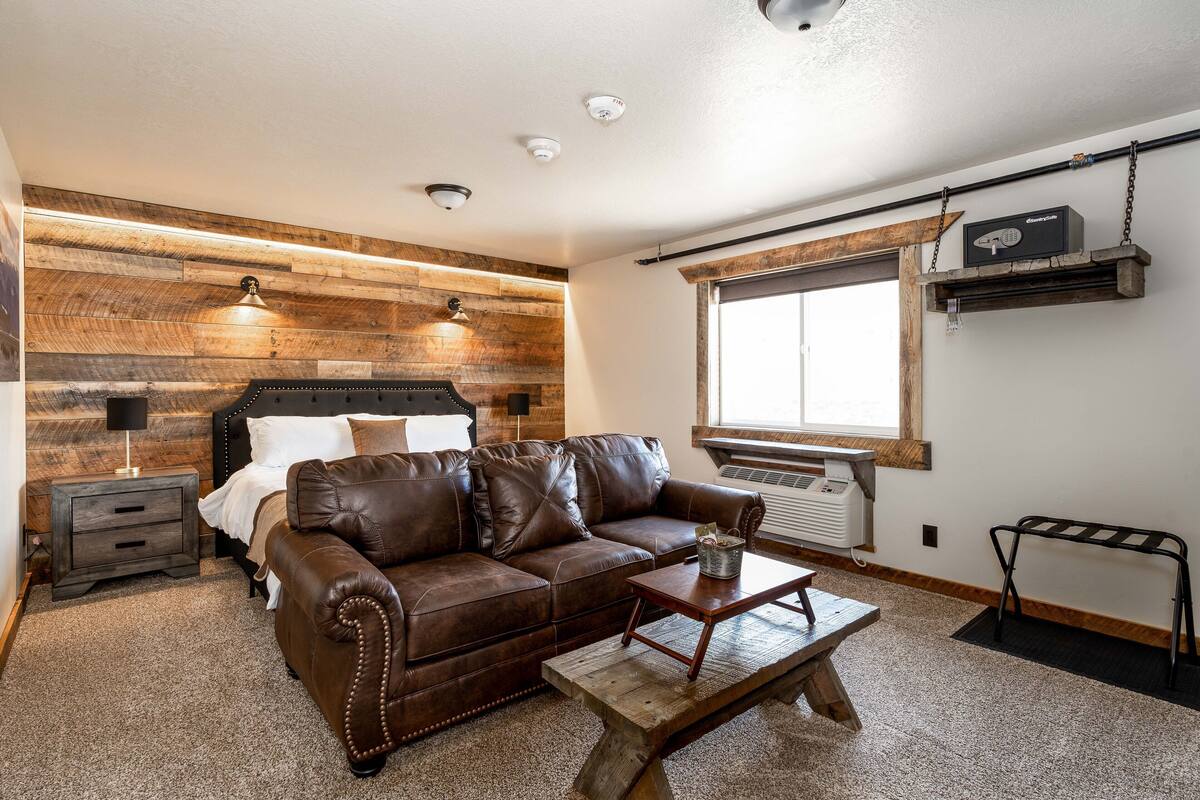 A bedroom with a brown leather sofa, a wooden coffee table, a nightstand, and a bed with a headboard.
