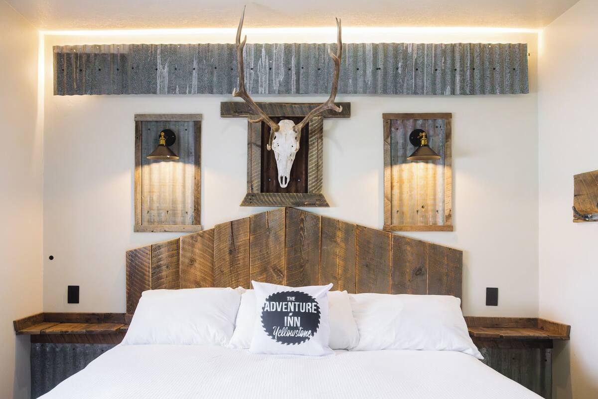 A bed with a wooden headboard, a wall-mounted deer skull, and two wall sconces.