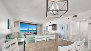 A spacious room with a large wooden dining table, white chairs, a chandelier, a TV, and a view of the ocean. - Gulf View Balcony ~ Seasonally Heated Pool (Miramar Beach)