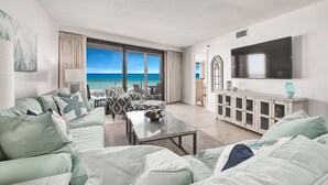 A living room with a large window overlooking the sea, a sofa set, a coffee table, and a TV mounted on the wall. - Gulf View Balcony ~ Seasonally Heated Pool (Miramar Beach)