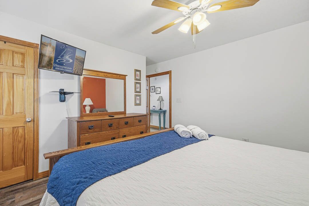 A bedroom with a bed, wooden dresser, ceiling fan, and a framed picture on the wall.