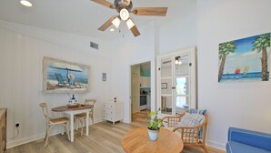 Un coin repas avec une table en bois, des chaises blanches et un ventilateur de plafond. Un tableau représentant une scène de plage est accroché au mur.
