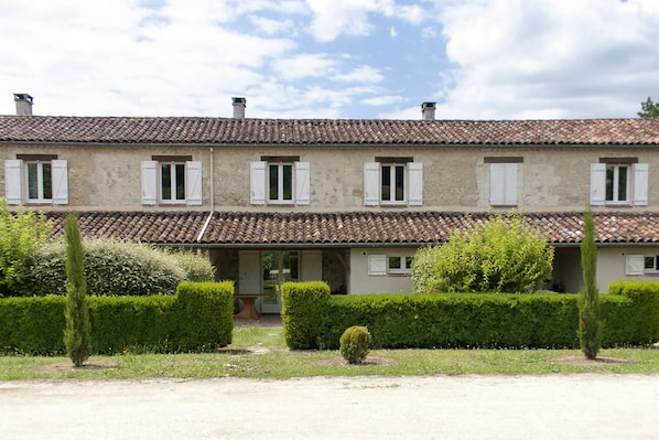A two-story building with a tiled roof, white shutters, and a well-maintained garden with hedges and a small tree.