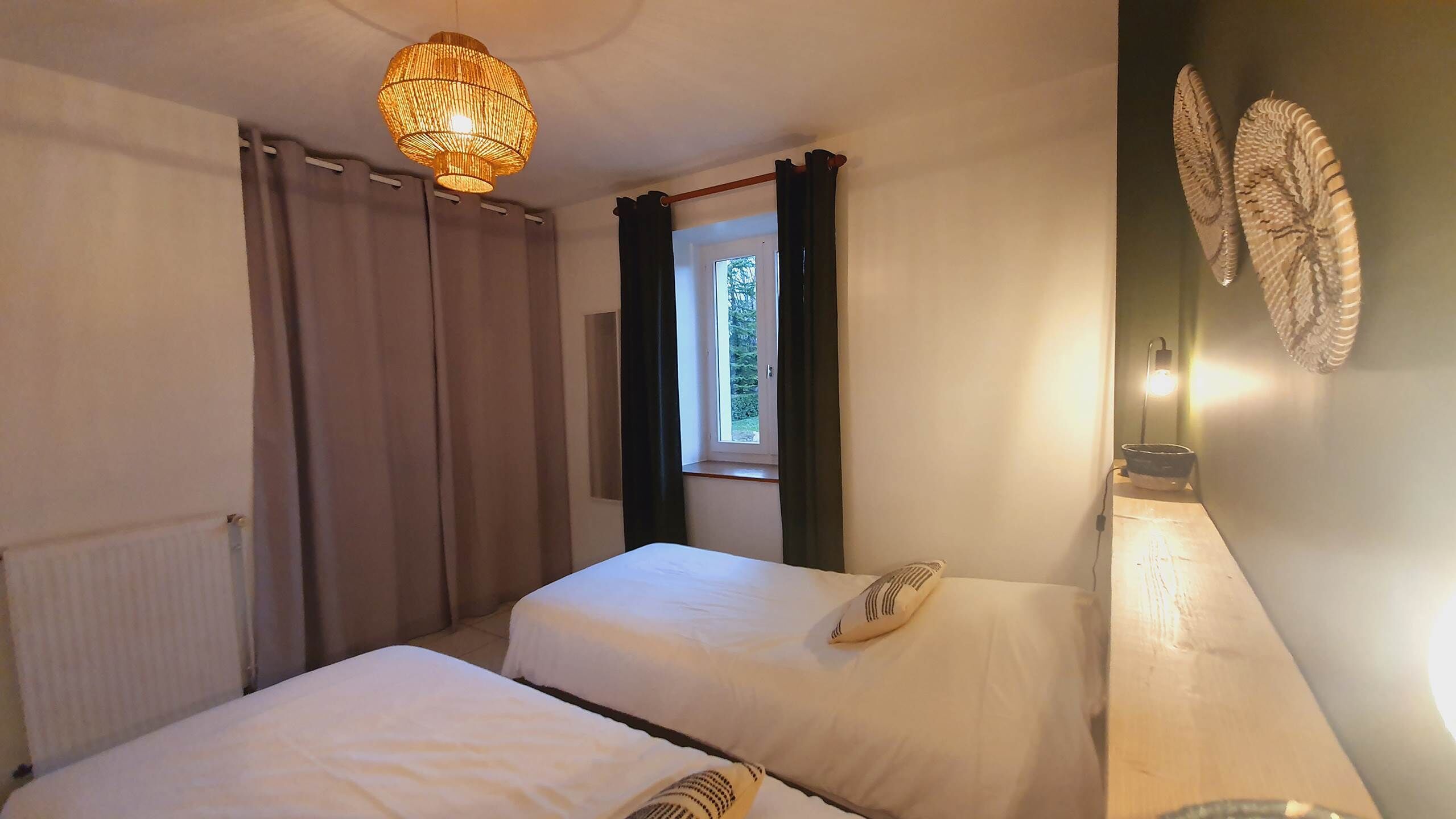 A room with two beds, a window with curtains, a wall-mounted light, and a decorative wall piece.