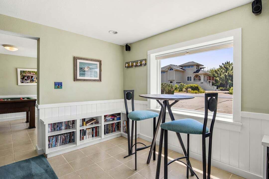 A room with a pool table, bar stools, a bookshelf, and a window with a view of houses.