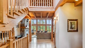 A wooden staircase leading to a lofted area with a stone fireplace, a large window with a view of greenery, and a framed picture on the wall. - Stunning 5BR Family Lodge—Hot Tub, Huge Deck & Mountain Views (Silverthorne)